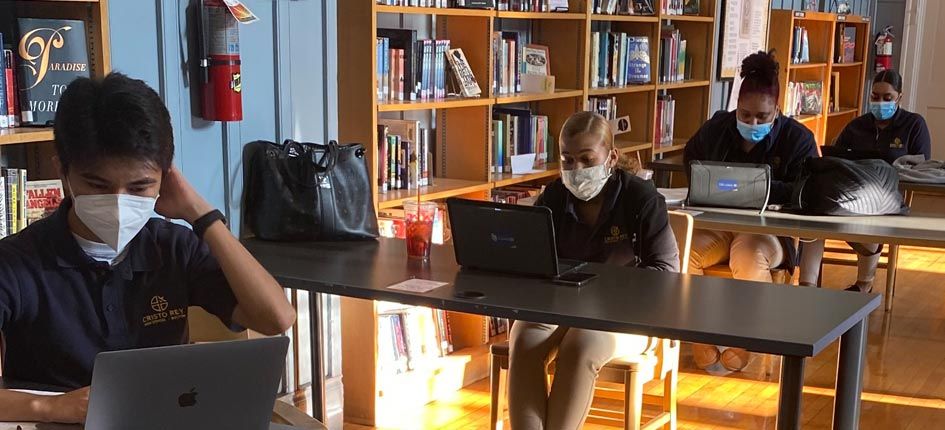 Four students, one boy and three girls, studying in a library. Each is at their own desk, 6 feet apart, wearing masks and working on a laptop.